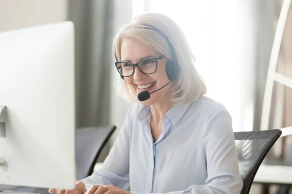 Happy old businesswoman in headset making call looking at computer — Stock Photo, Image