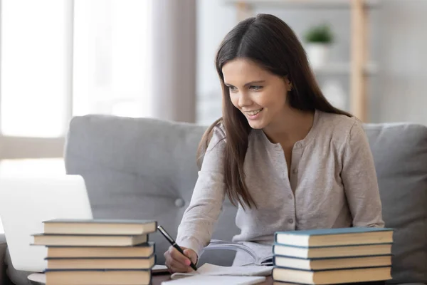 Chica estudiante positiva sentada en el sofá en la mesa y estudiando —  Fotos de Stock