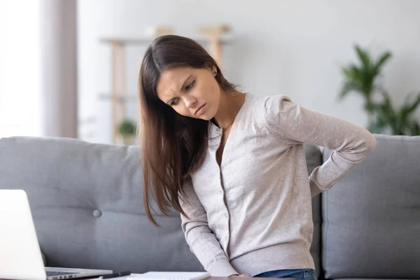 Нездоровая молодая женщина, сидящая на диване, страдает от болей в спине — стоковое фото