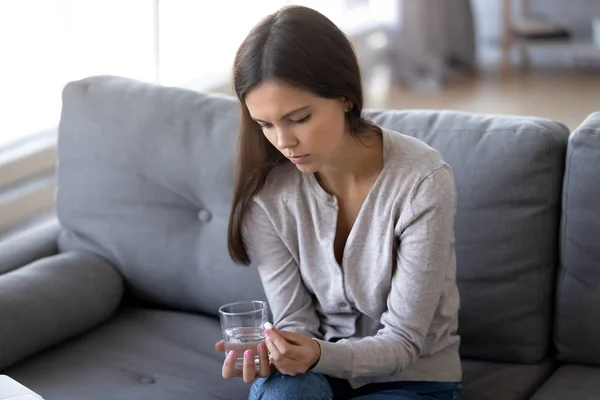 Грустная женщина сидит в помещении с таблетками и стаканом воды — стоковое фото