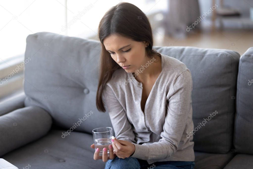 Sad female sitting indoors holding pill and glass of water