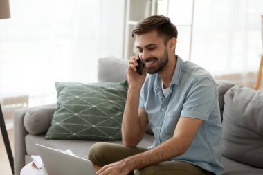 Mutlu adam laptop kullanarak gülümseyen ve evde telefonda konuşurken