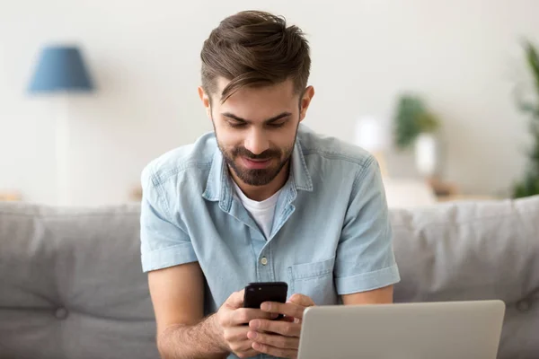 Smiling man sitting at home, using phone, chatting, typing message — Stock Photo, Image