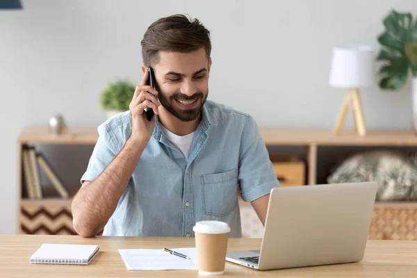 Uśmiechnięty zadowolony człowiek rozmawia na telefon i za pomocą laptopa w miejscu pracy — Zdjęcie stockowe
