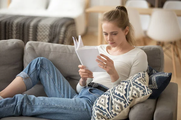 Молодая женщина сидит на удобном диване и читает бумажную книгу в одиночестве — стоковое фото
