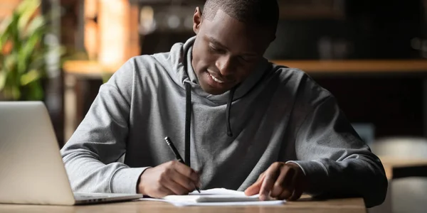 Сосредоточенный африканский студент сидит в помещении и пишет на блокноте — стоковое фото