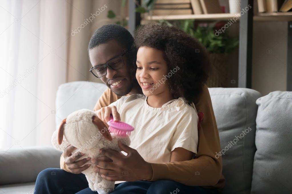 Biracial daughter sitting on daddy lap combing stuffed toy dog
