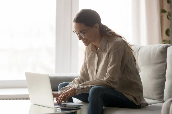 Mujer en gafas sentada en el sofá sonriendo escribiendo en el ordenador — Foto de Stock