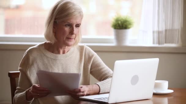 Пожилая женщина, держащая счета, использует ноутбук, чтобы проверить онлайн платежи — стоковое видео