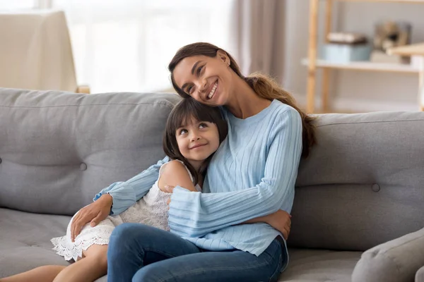 Мила щаслива дочка обіймає молоду маму, сидячи на дивані — стокове фото