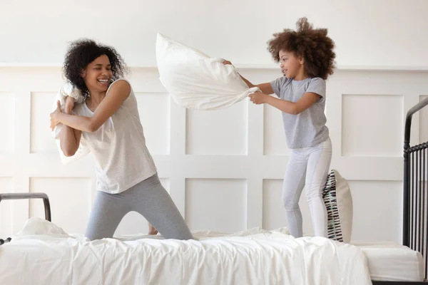 Счастливые африканские мама и дочь дерутся подушками на кровати — стоковое фото