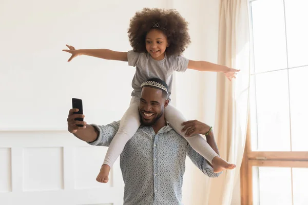 Feliz africano papá usando corona piggybacking pequeño hija tomando selfie —  Fotos de Stock