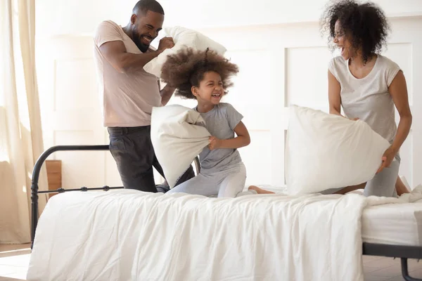 Carefree black parents small kid enjoying pillow fight on bed