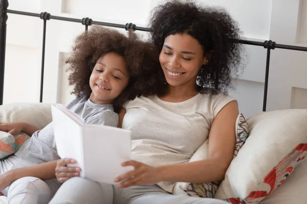 Feliz madre africana leyendo libro relajándose en la cama con su hija — Foto de Stock