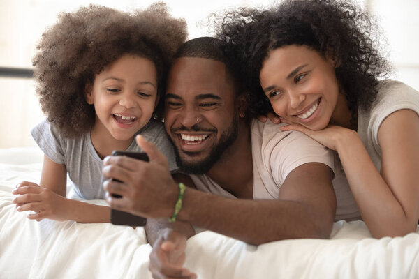 Cheerful african parents and kid laughing using smartphone in bed