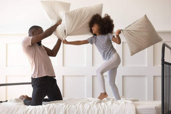 Cute daughter playing pillow fight on bed with african dad