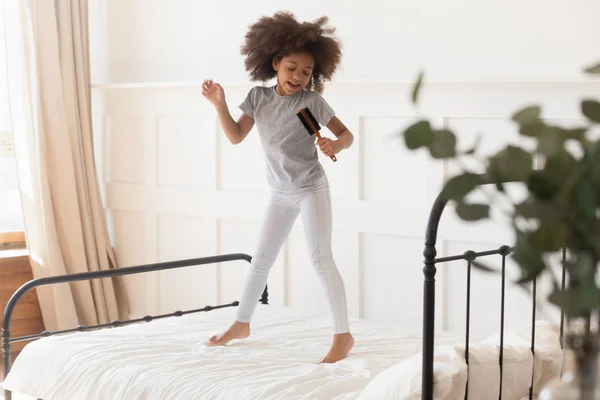 Divertente ragazza africana che salta sul letto cantando in spazzola per capelli — Foto Stock