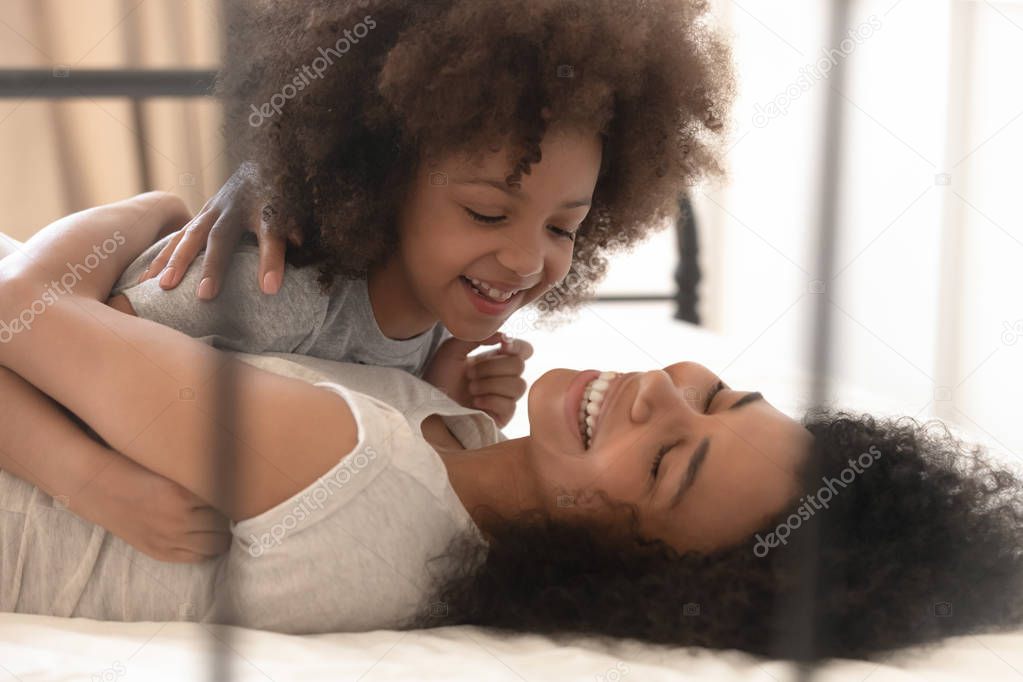 Happy mixed-race kid daughter and mum playing lying on bed