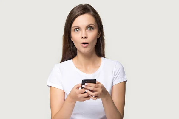 Amazed woman holding smartphone looking at camera posing on grey