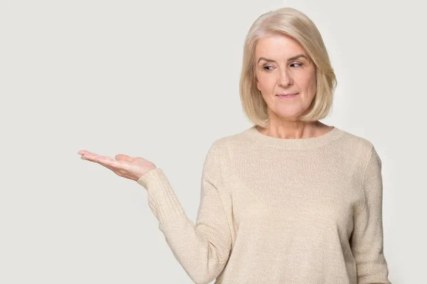 Зрелая женщина показывает открытую ладонь предлагая новый продукт серый фон — стоковое фото