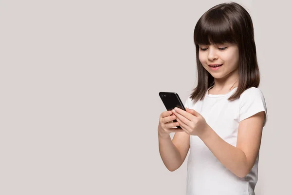 Mała dziewczynka za pomocą smartfona stojący na bok na szarym tle — Zdjęcie stockowe