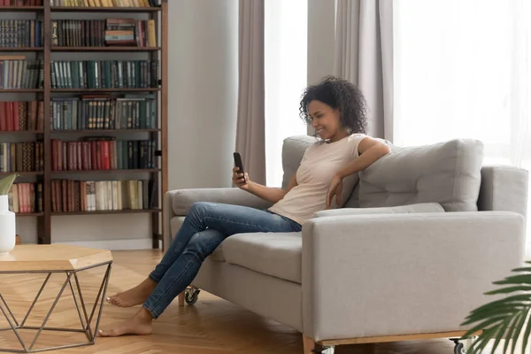 Happy young african american woman using smartphone sit on sofa — Stok Foto