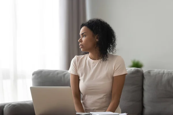 Uttråkad afrikansk flicka tittar bort sitta på soffan med laptop — Stockfoto