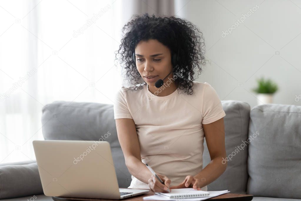 Young african woman wearing headset looking at laptop making notes