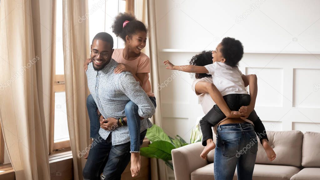 Happy african parents piggyback children laugh play in living room