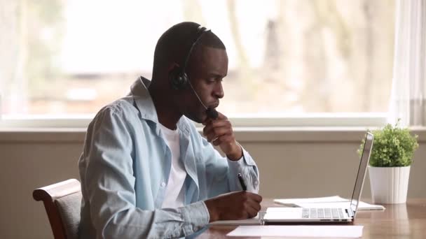 Homem de negócios africano focado usar fone de ouvido olhar para laptop fazer notas — Vídeo de Stock