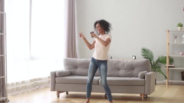 Mujer africana sosteniendo un smartphone bailando escuchando música en casa — Vídeos de Stock