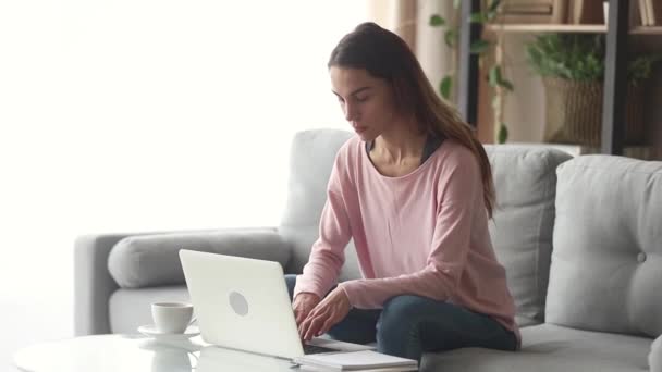 Mujer joven feliz seria usando el ordenador portátil sentado en el sofá — Vídeos de Stock