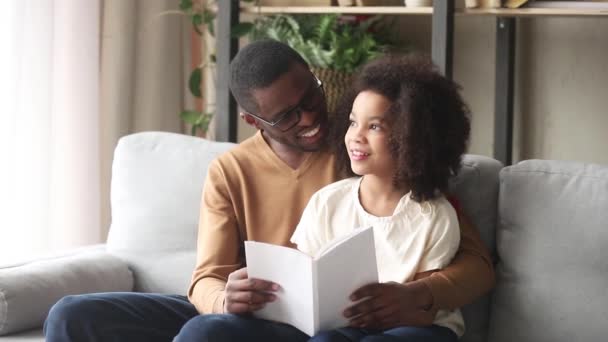 Любящий чёрный отец обнимает дочку, читает дома книгу. — стоковое видео