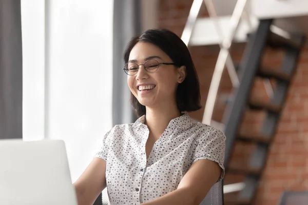 Alegre asiático mujer de negocios riendo mirando portátil sentado en escritorio — Foto de Stock