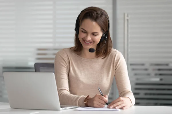 Businesswoman receptionist wear wireless headset looking at laptop make notes — Stock Photo, Image