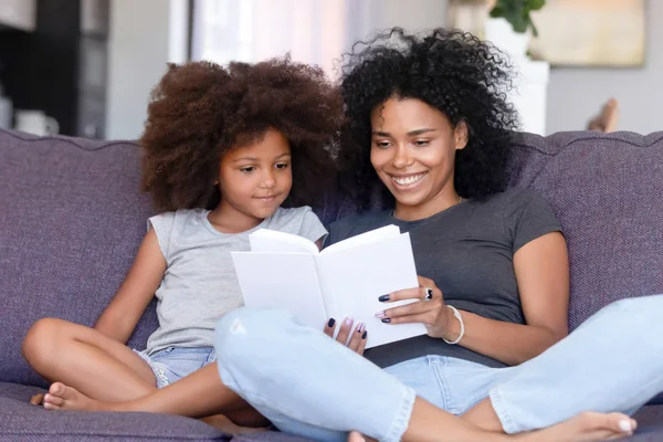 Madre africana e hija pequeña sentadas en el sofá leyendo libro — Foto de Stock