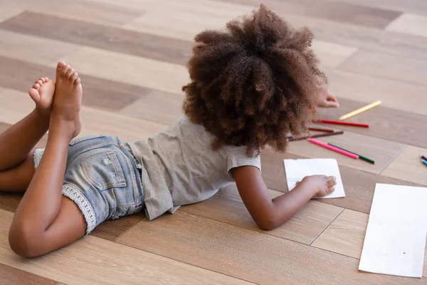 Back view african little girl drawing lying on warm floor