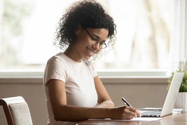 African woman in glasses sitting near computer holding pen writing — Stock Photo, Image