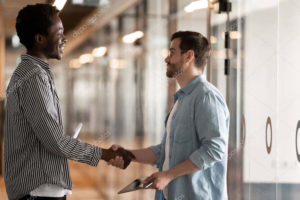 Two diverse male colleagues greeting handshaking standing in office hall