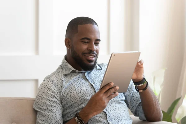 Smiling african man using digital tablet reading e-book at home