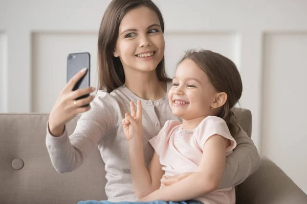 Cheerful funny mother holds smartphone take selfie with daughter