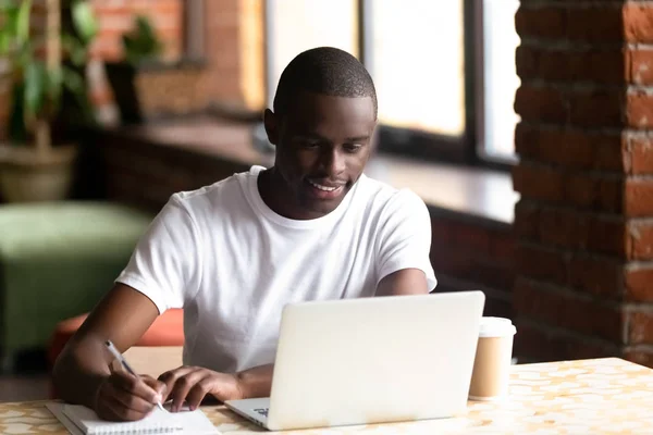 Sorridente maschio nero che studia sul computer portatile prendendo appunti nel notebook — Foto Stock