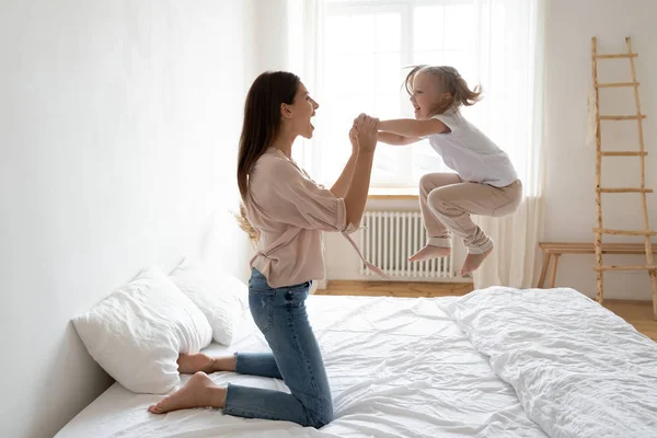 Pequeña hija frolics saltar en la cama sosteniendo las manos de madres —  Fotos de Stock