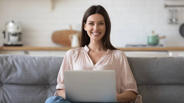 Женщина, сидящая на диване, держит компьютер и смотрит в камеру — стоковое фото