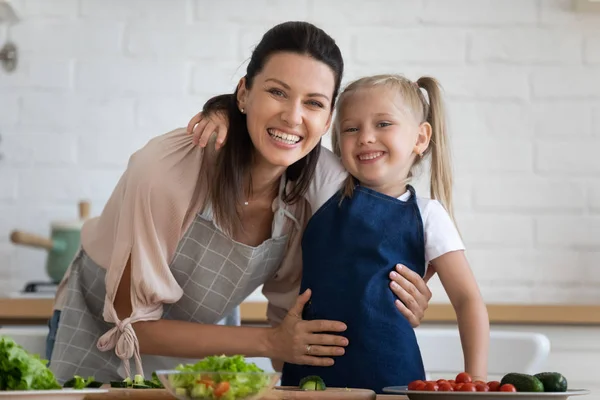 Moeder omarmt dochter kijken naar camera koken samen in de keuken — Stockfoto