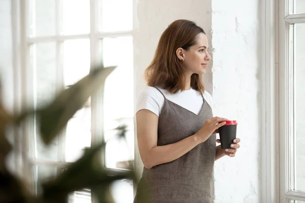 Vacker ung kvinna som håller kopp kaffe, ser ut fönster — Stockfoto