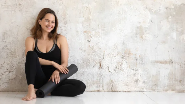 Feliz mujer sonriente sosteniendo alfombra de yoga negro, sentado en el suelo —  Fotos de Stock