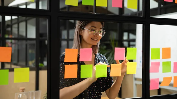 Sorridente asiatico businesswoman scrivere compiti su appiccicoso note su vetro parete — Foto Stock