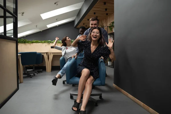 Happy funny diverse coworkers riding on chairs in modern office — Stock Photo, Image