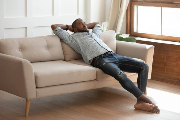 Feliz millennial descalço preto cara daydreaming no sofá . — Fotografia de Stock